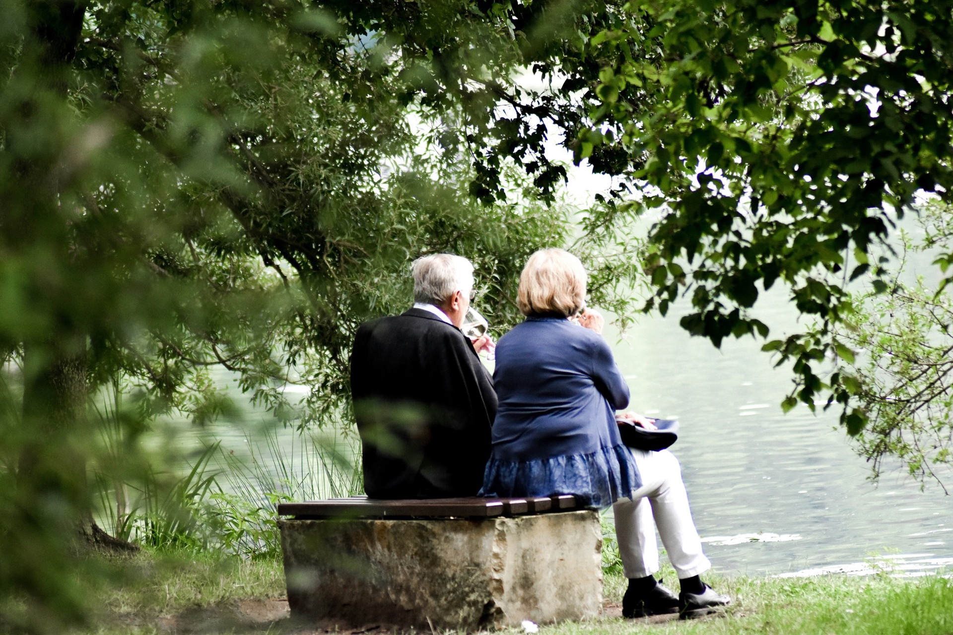 An elderly couple sitting together by a lake, framed by trees